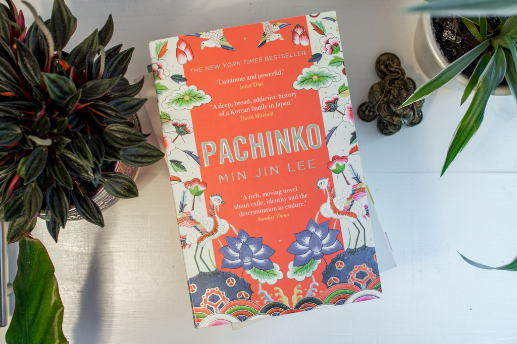 Top down view of the book cover Pachinko by Min Jin Lee next to two potted plants.