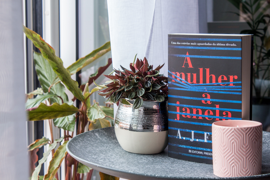 Photo of the book The Woman in the Window by A J Finn standing up right on a table next to a window.