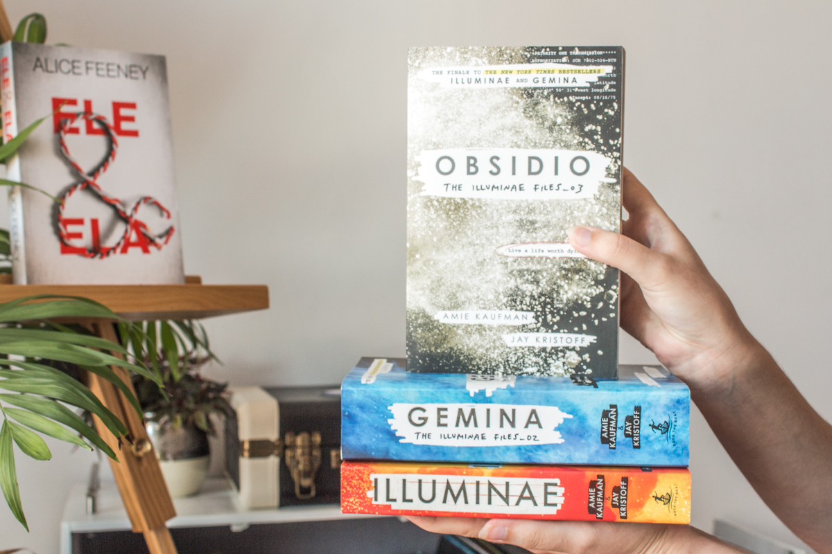 Holding a stack of the three books from the Illuminae files series with His & Hers out of focus in the background.