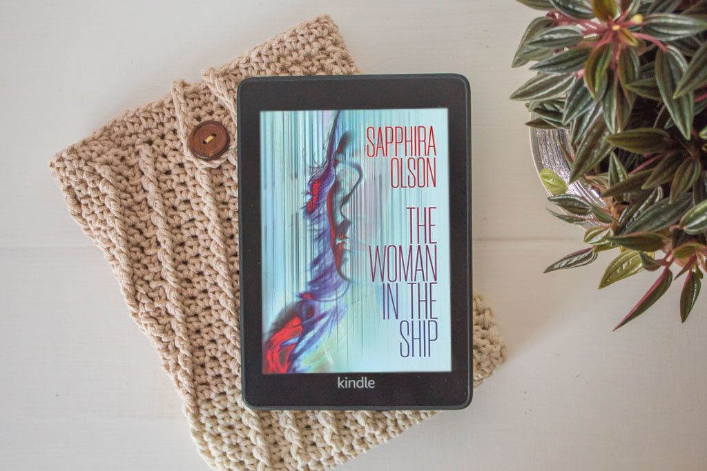 Kindle lying on a table showing the coloured cover of The Woman in the Ship.