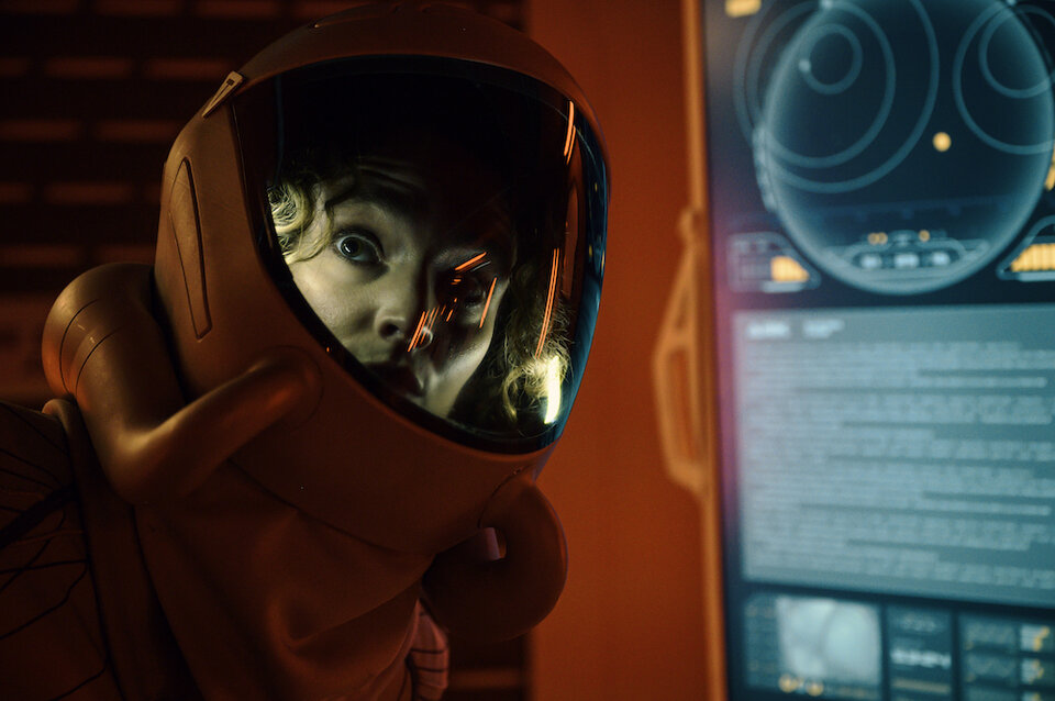 Astronaut in a spacesuit illuminated by the lights in the helmet next to a status screen.