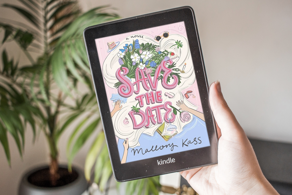 Holding a kindle showing the coloured cover of the book with a plant in the background.